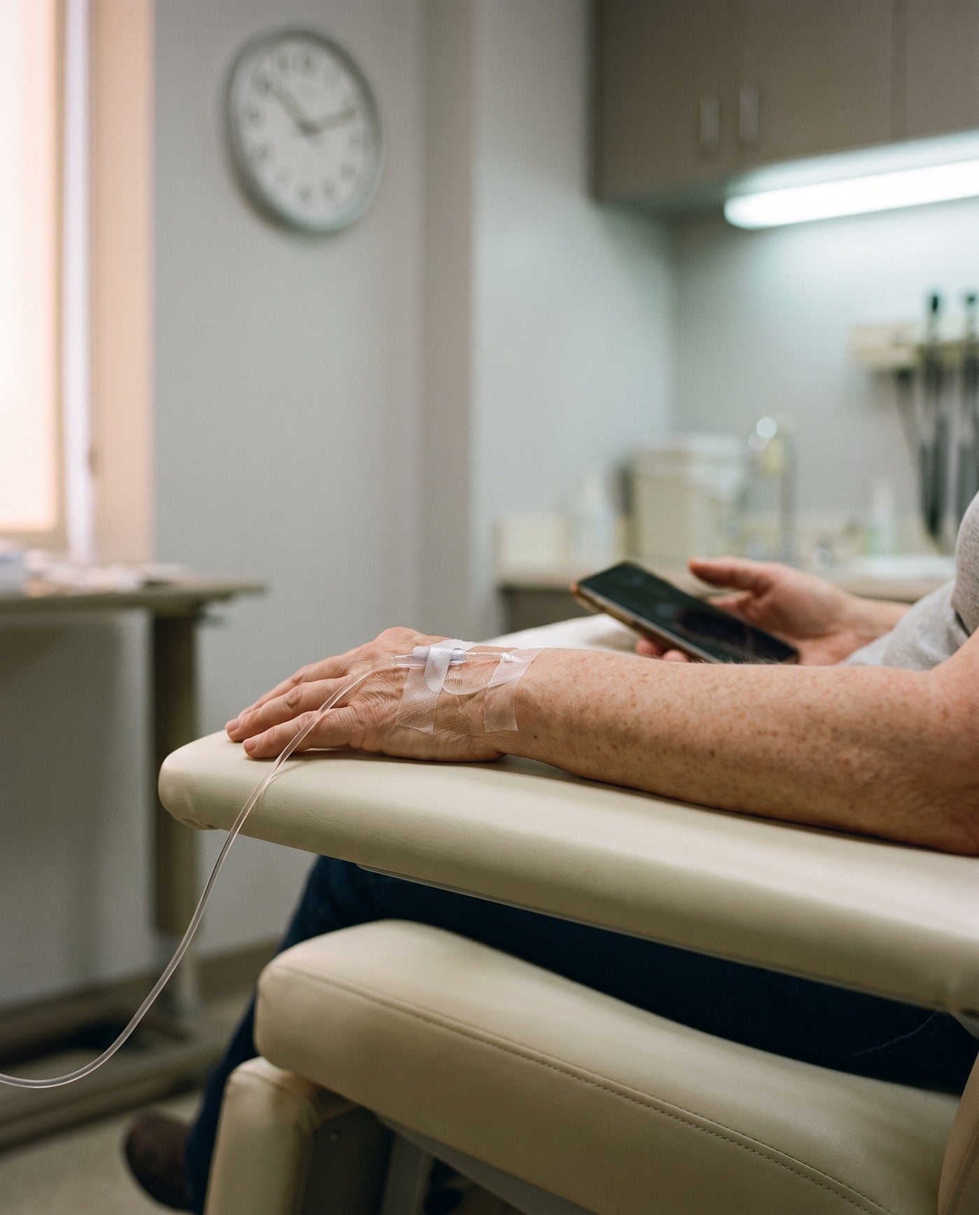 A woman's forearm resting on a clinic chair with a taped cannula and tubing, a wall clock out of focus behind