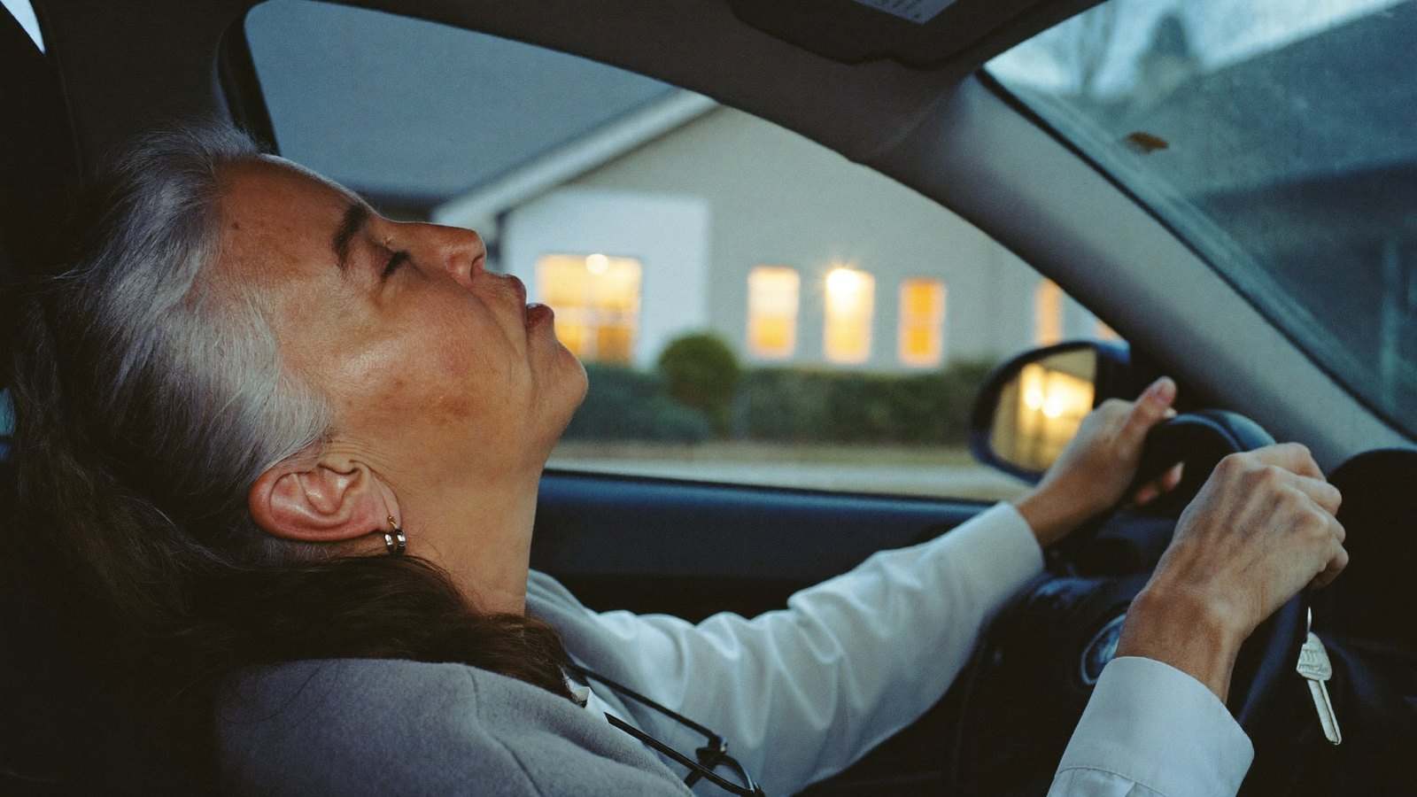 Woman in her fifties sitting in her parked car outside her house at dusk